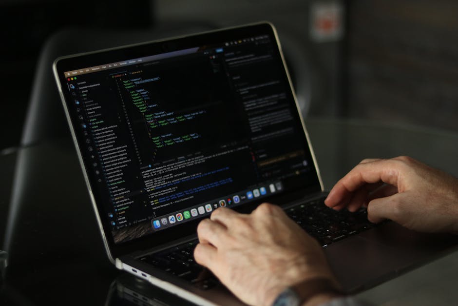 A person using a laptop outdoors, with their hands visible typing on the keyboard. The screen displays a dark-themed coding interface with colorful lines of code and text, set on a sleek, modern lapto
