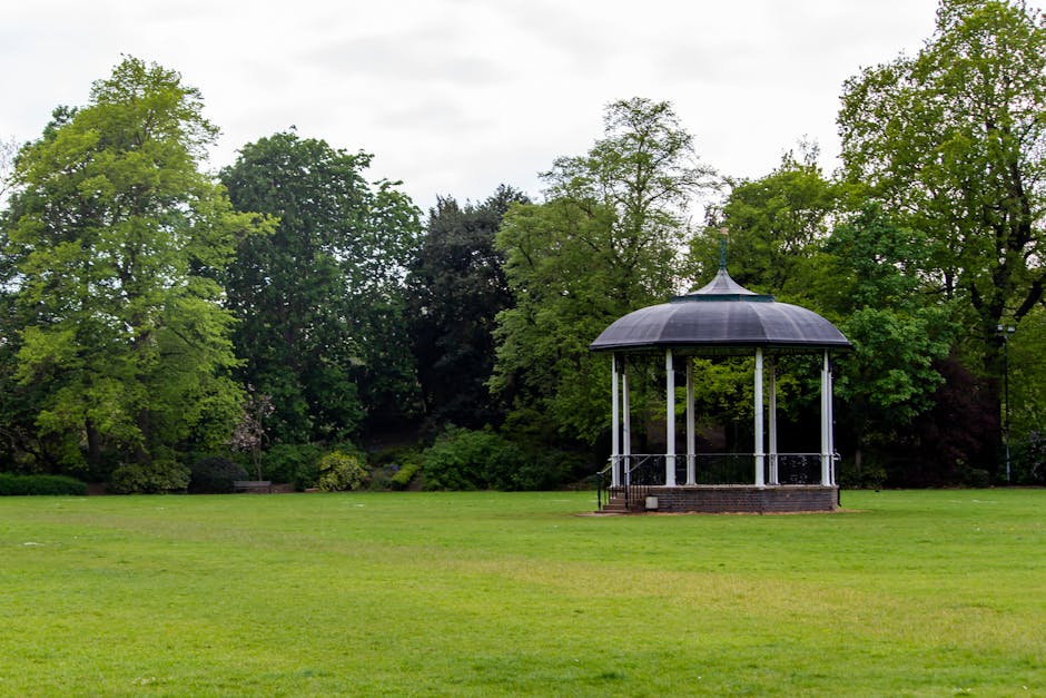 A spacious, well-maintained park with a lush, green lawn and a circular bandstand structure with a dark, domed roof and white support columns situated centrally. The grassy area is evenly trimmed with