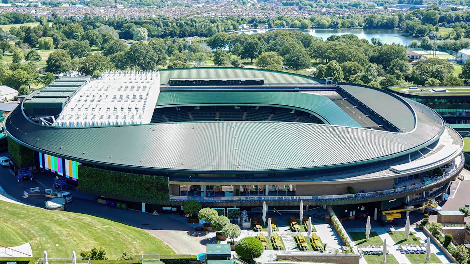 A groundskeeper dressed in dark clothing and a cap is pushing a lawn mower across the grass of a large sports stadium, which appears to be a tennis or football arena with tiered seating and floodlight